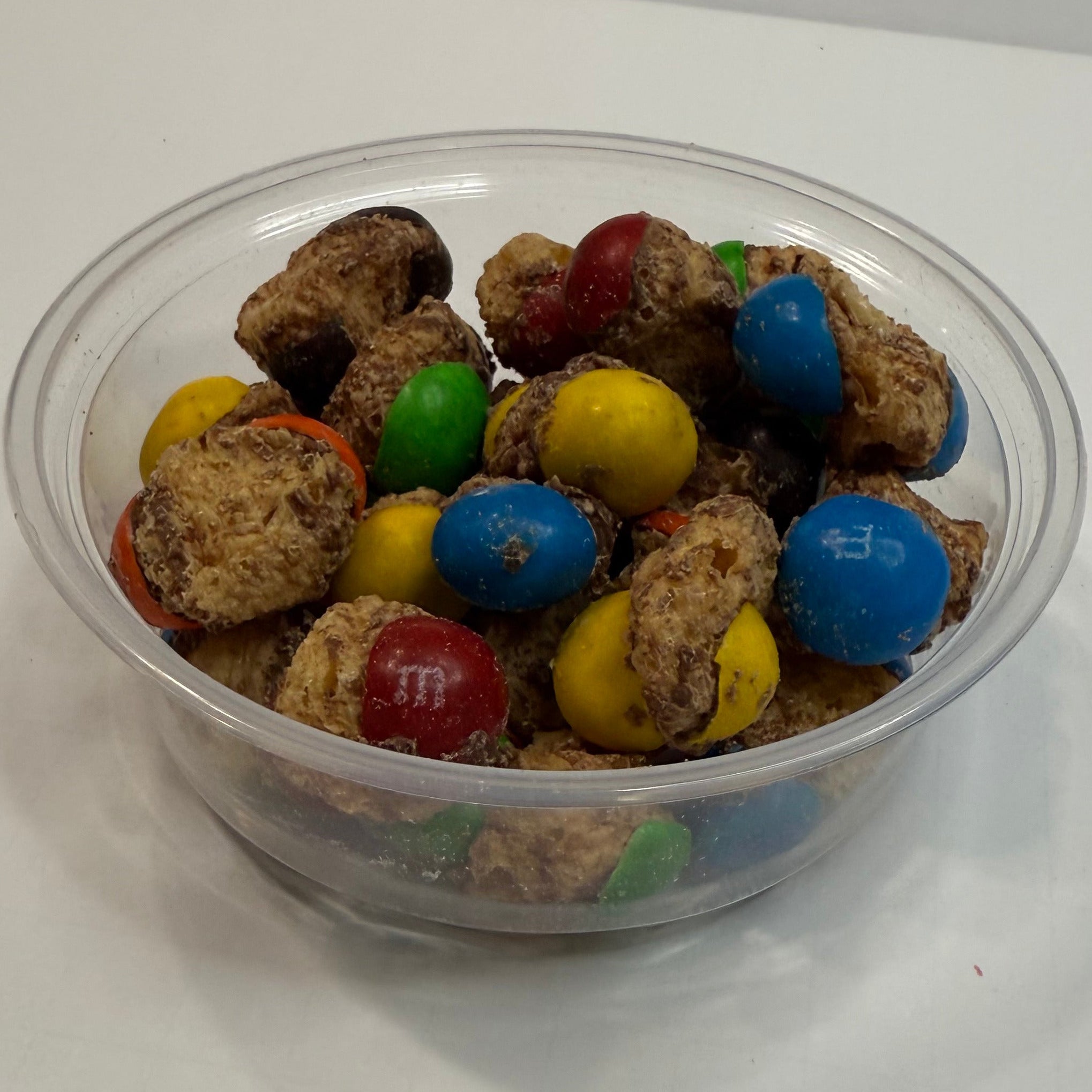 Clear plastic container with colorful freeze dried caramel M&Ms on a white surface
