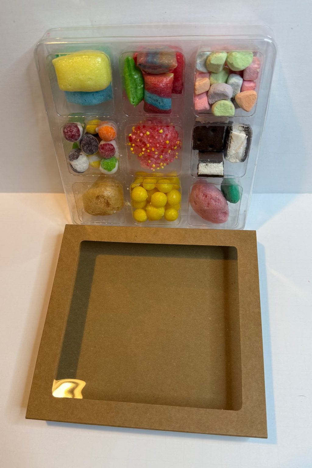 Assorted colorful candies in a transparent plastic container on a white surface with a brown cardboard box.
