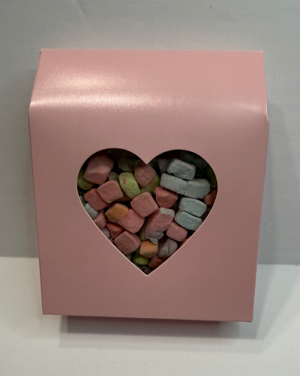 Pink box with a heart-shaped window displaying colorful freeze dried marshmallow charms on a white background