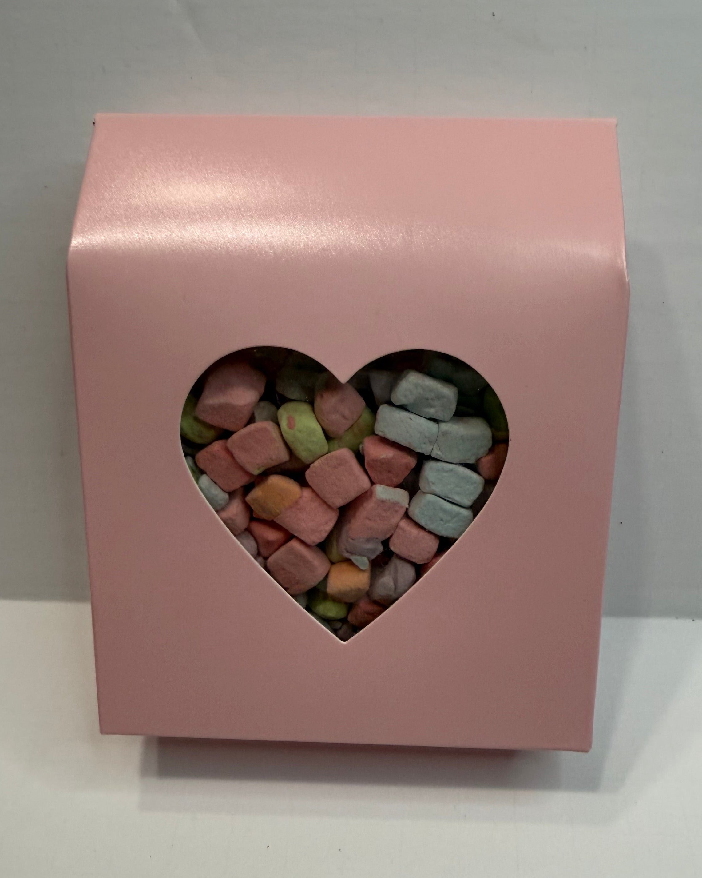 Pink box with a heart-shaped window displaying colorful freeze dried marshmallow charms on a white background