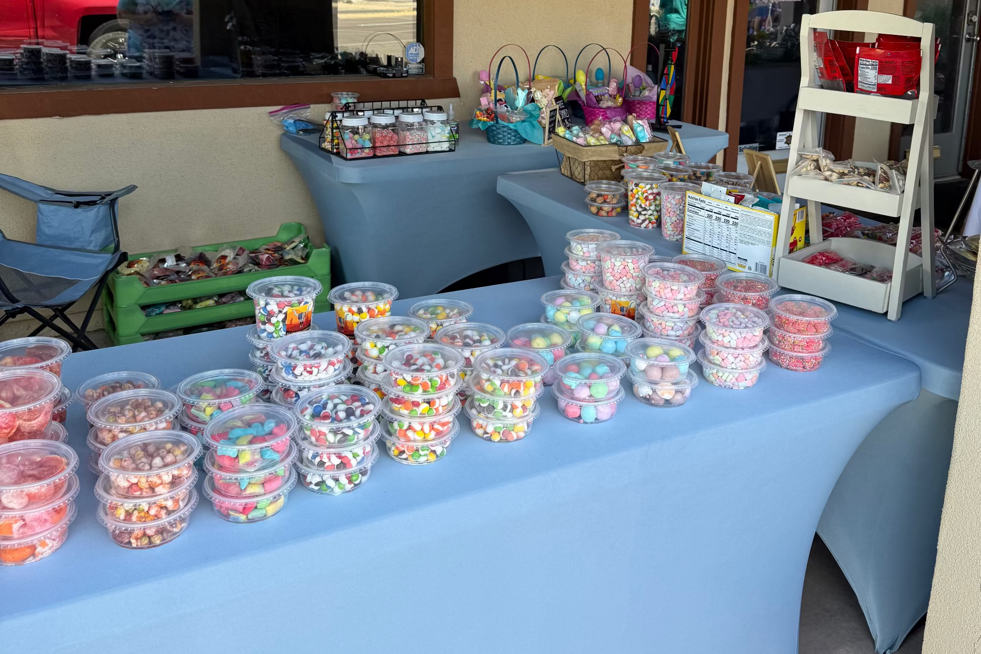 Table with containers of freeze dried candy on a blue tablecloth, with chairs and a cart in the background.