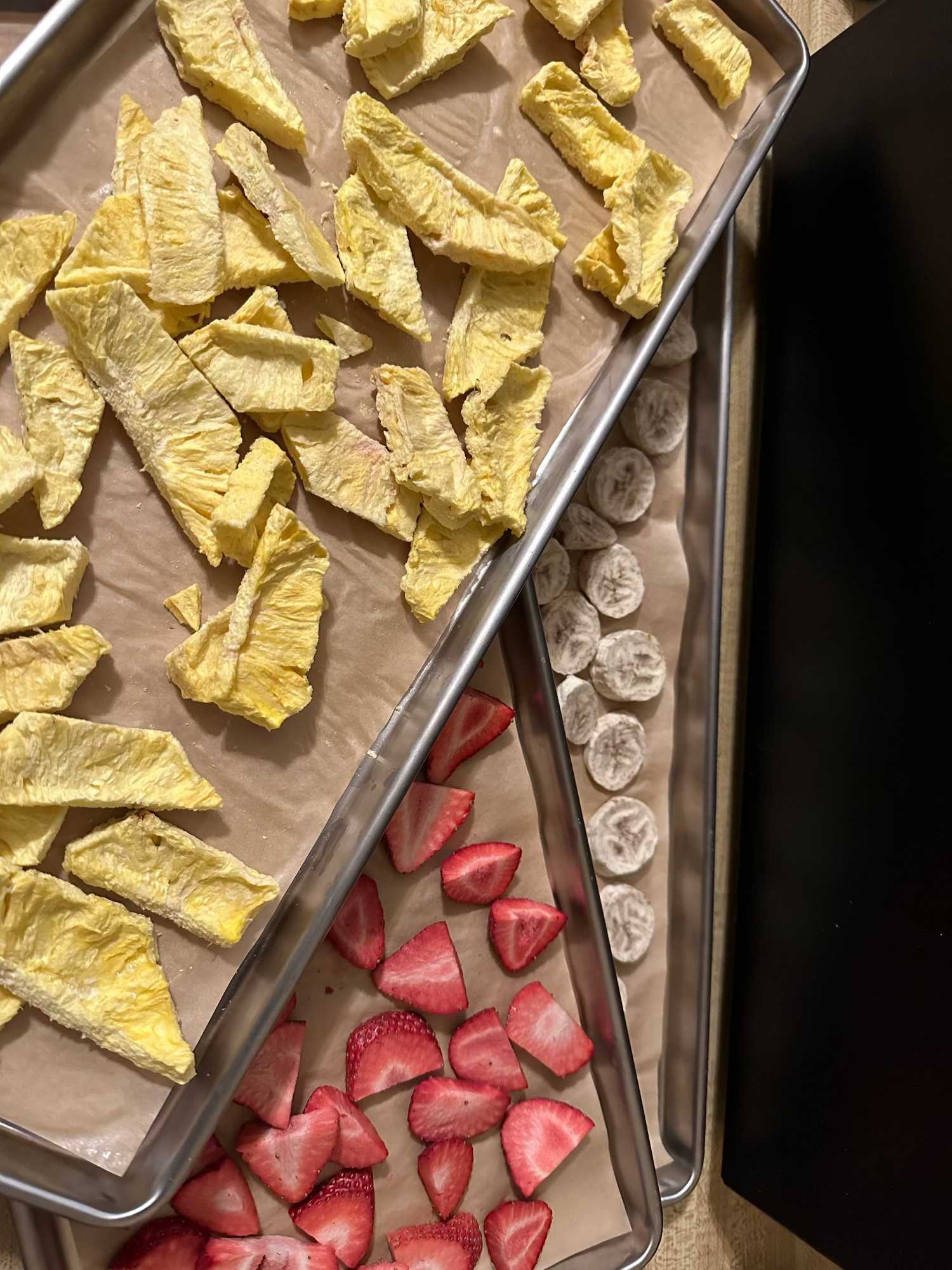 Three baking trays with freeze dried sliced strawberries, banana slices, and yellow vegetable pieces on a dark surface.